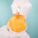 A gloved hand pours orange Carnival King snow cone syrup into a cup.