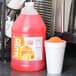 A jug of Carnival King Orange Slushy concentrate next to a cup of slush.