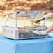 A woman uses a Carnival King glass sneeze guard to put food on a hot dog roller grill.