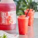 A plastic cup with a red slushy drink and a straw sitting next to a pitcher of red liquid.